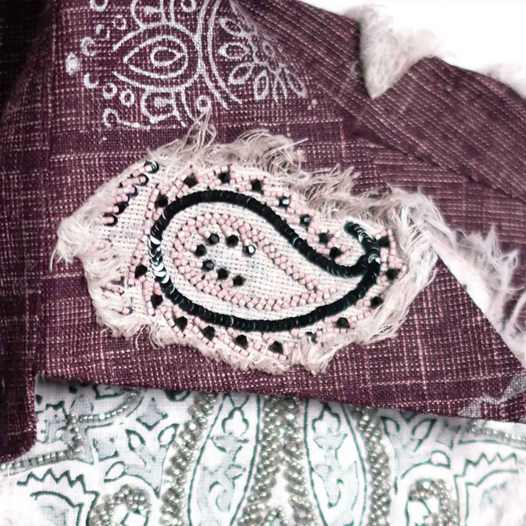 A close-up of a textured mauve-brown woven fabric with a large embroidered paisley patch in light pink, black, and white, layered over a white patterned textile.