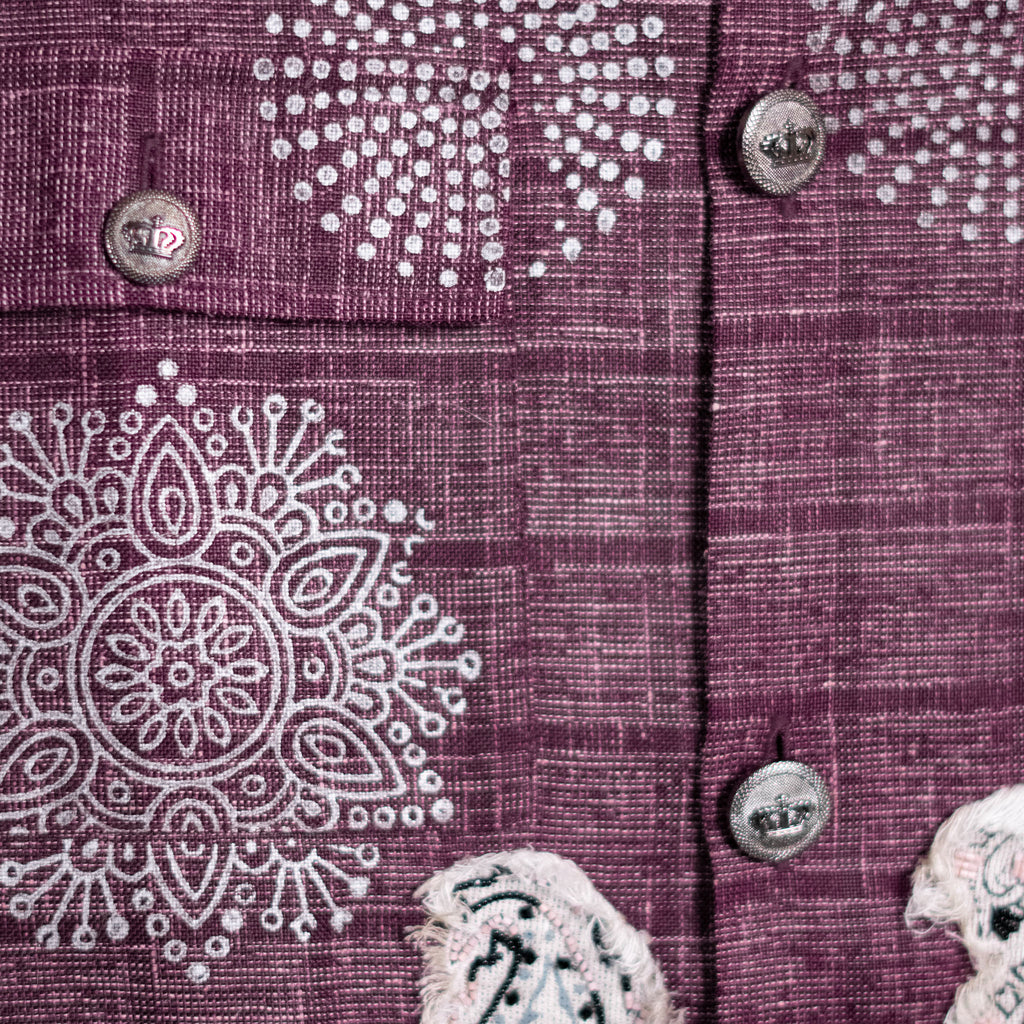 Close-up of a textured purple woven fabric garment with white embroidered geometric and floral patterns, featuring silver-toned buttons.