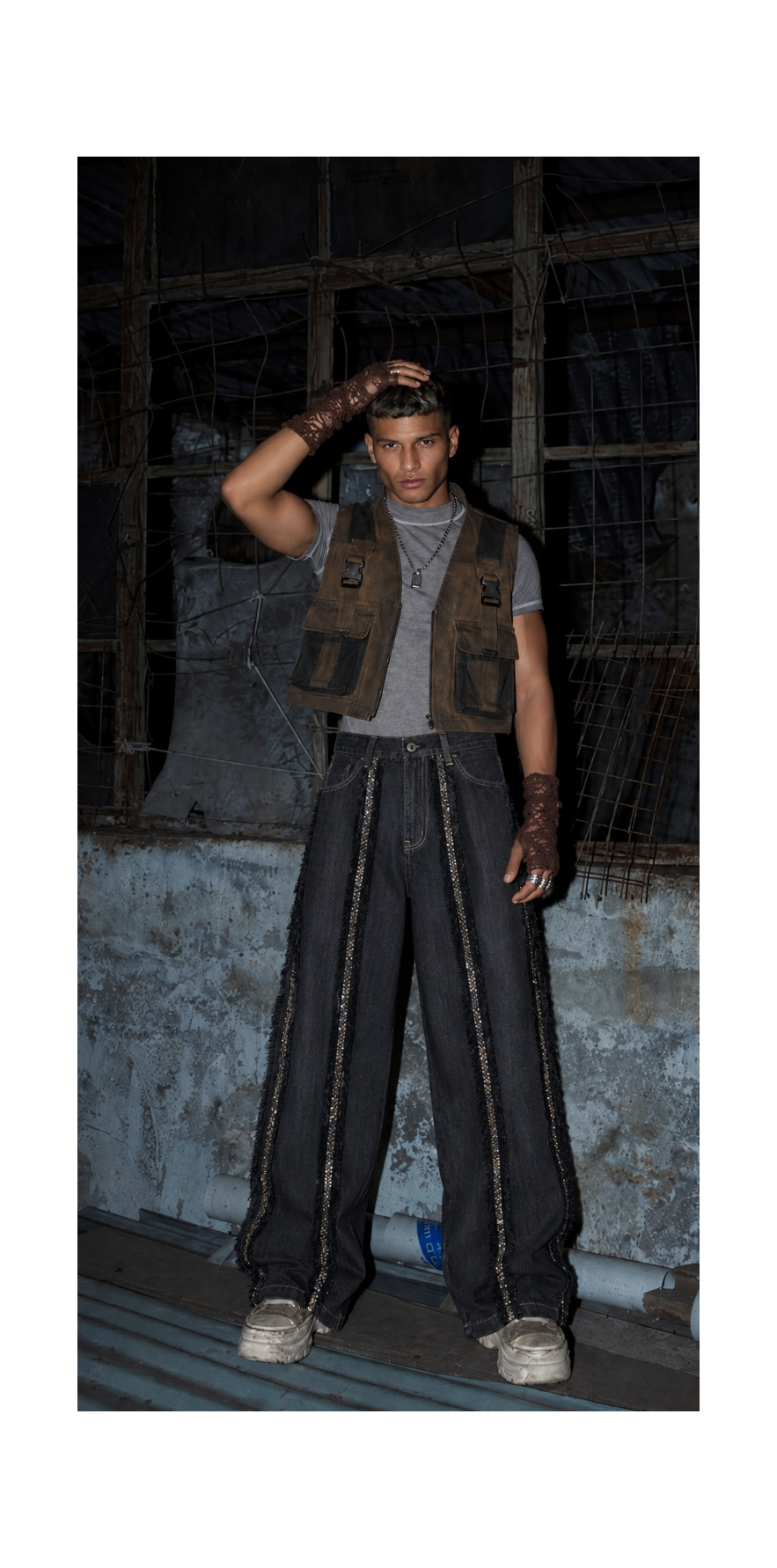 A model stands in a gritty, industrial setting wearing a brown utility vest over a gray t-shirt, dark wide-leg pants with chain details, and white sneakers.