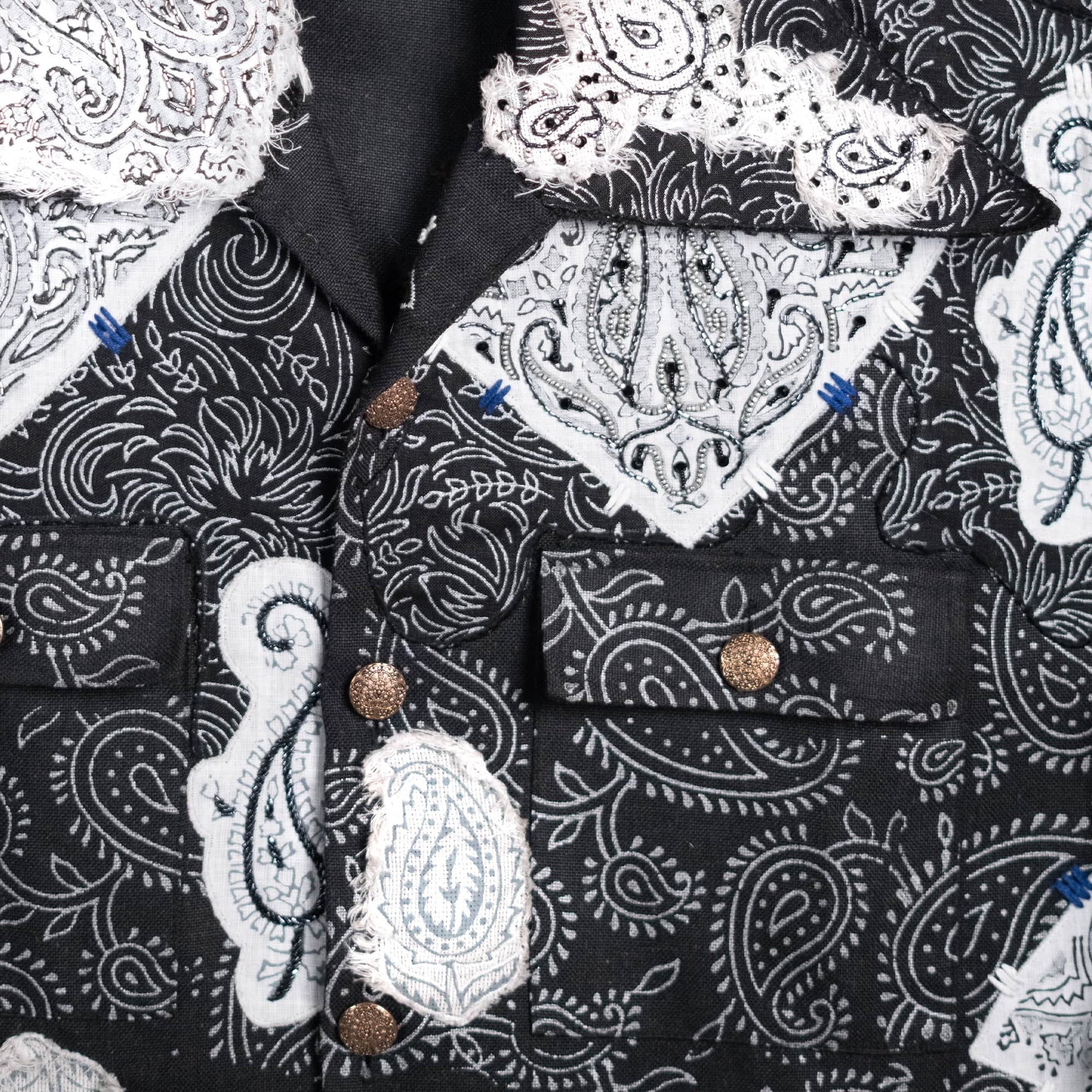 Close-up of a black button-front garment with a white paisley bandana print and white lace applique patches, plus bronze-toned buttons.