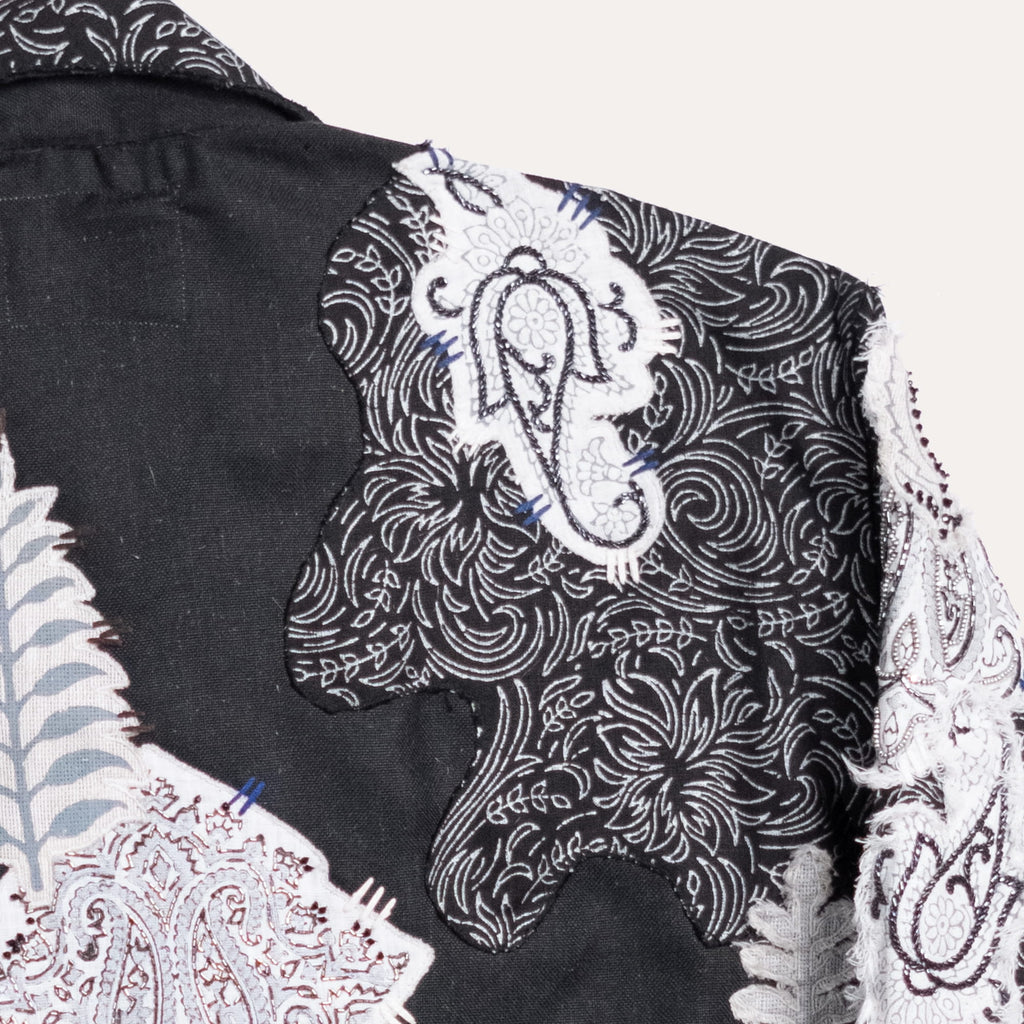 Close-up of a black fabric garment with ornate white and gray embroidered paisley and floral patterns, plus a small light blue accent, shown against a plain light background.