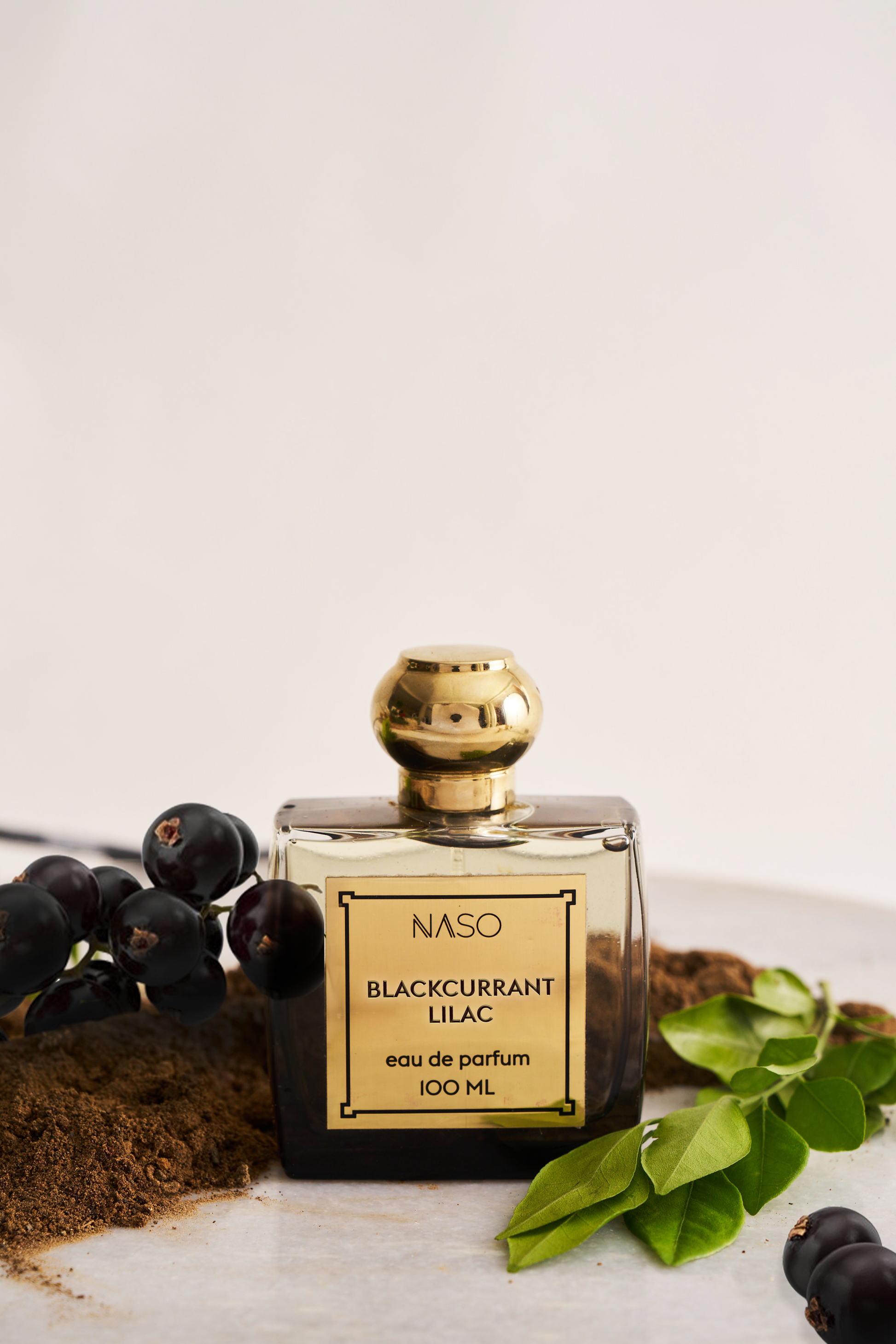 A 100 ml bottle of NASO Blackcurrant Lilac Eau de Parfum with a gold cap and gold label, styled on a marble surface with blackcurrants, green leaves, and coffee powder against a warm off-white background.