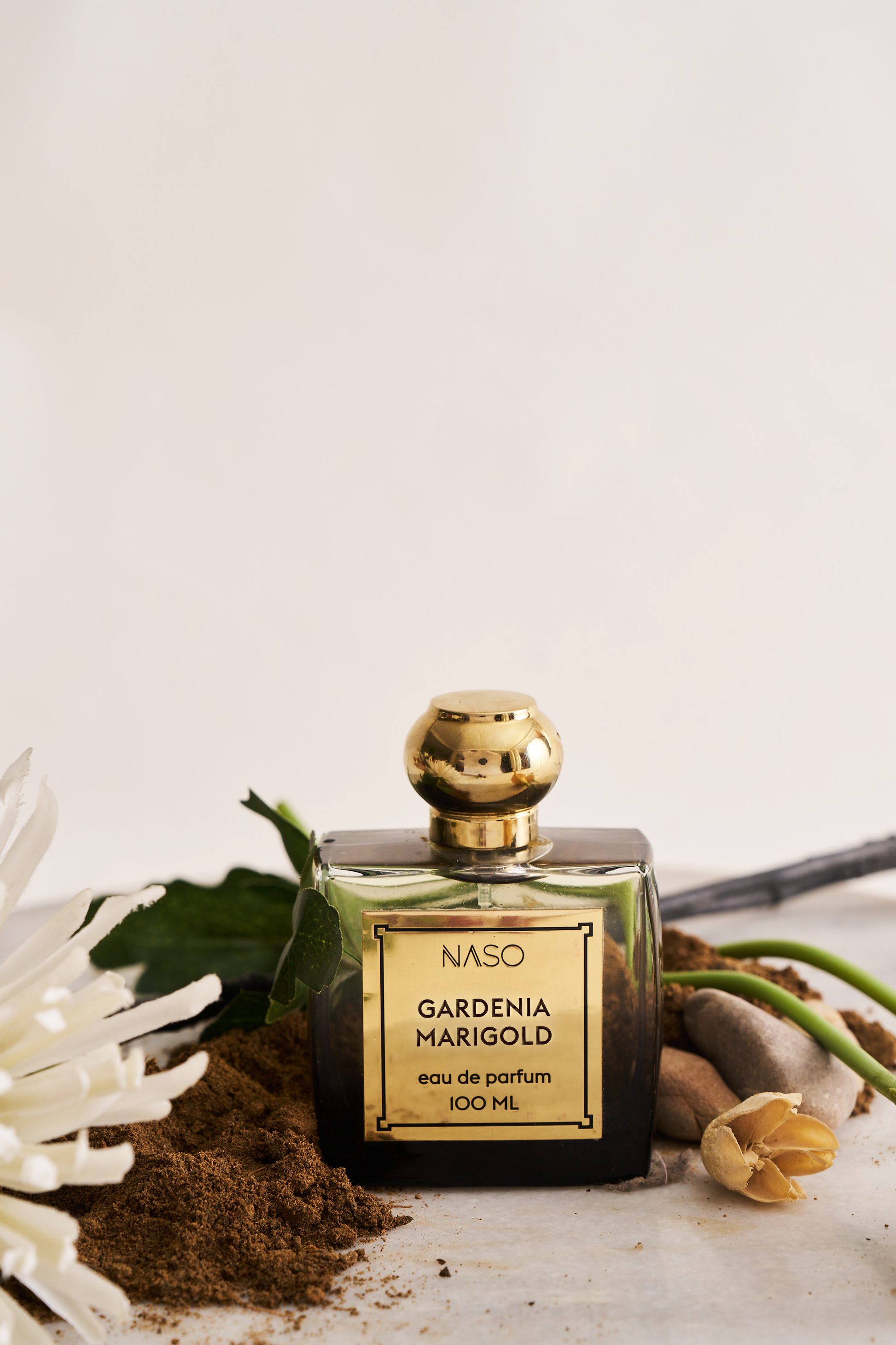 A 100ml bottle of NASO Gardenia Marigold Eau de Parfum, featuring a dark glass body, a gold cap, and a gold label. The bottle is styled on a neutral surface with gardenia petals, white chrysanthemums, and soil against a clean, off-white background.