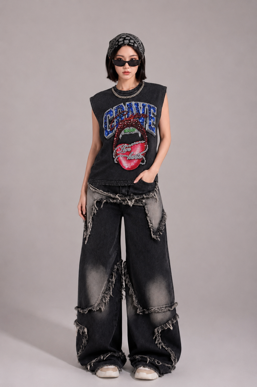 A model stands against a neutral gray studio background wearing a black sleeveless graphic top with a colorful tongue-and-lips design, distressed black wide-leg jeans with frayed details, a black beanie, and dark sunglasses.