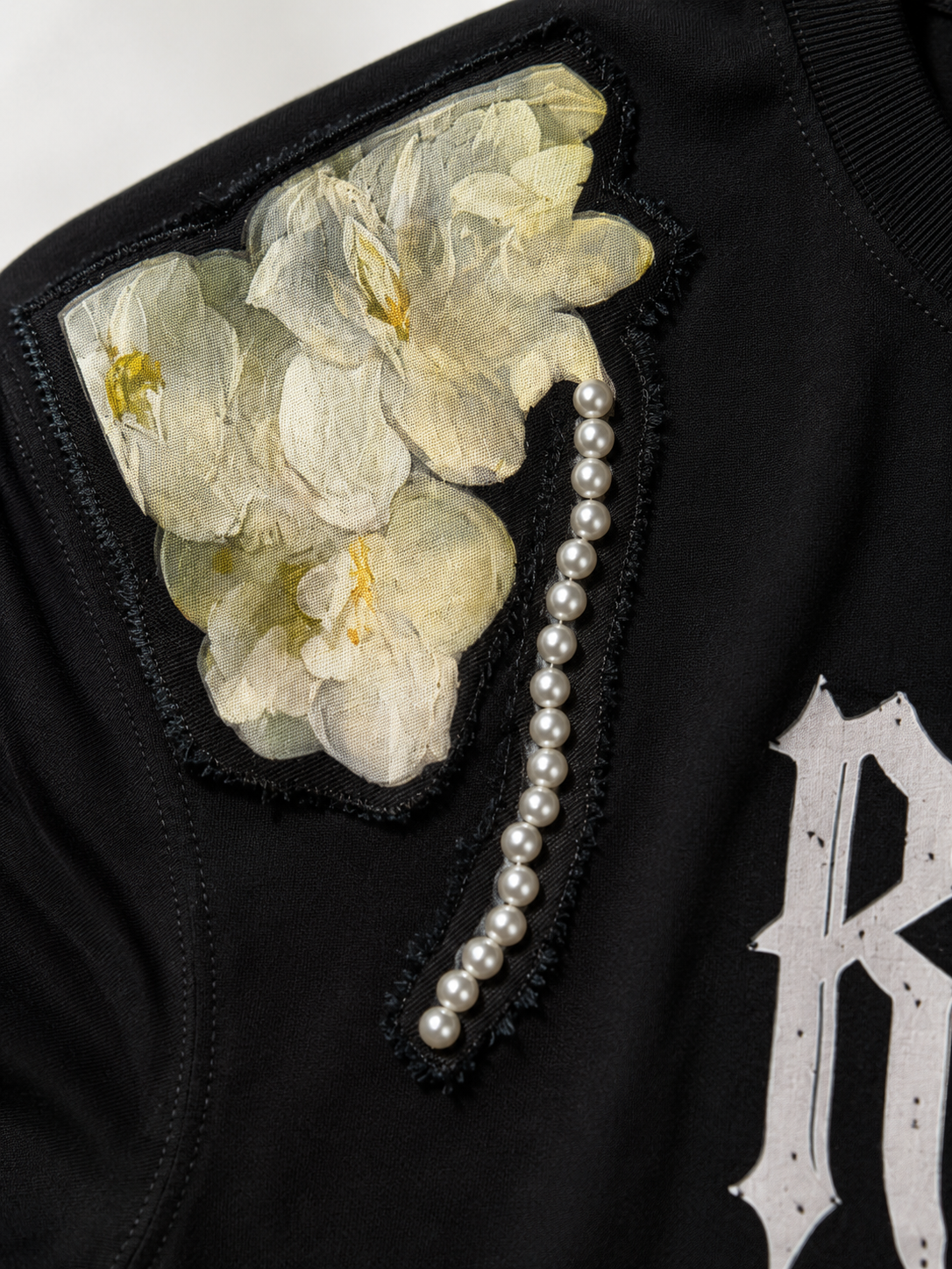 Close-up of a black garment featuring a large floral applique patch in pale cream tones, a vertical strand of white pearls, and a distressed white letter 'R' on the chest.