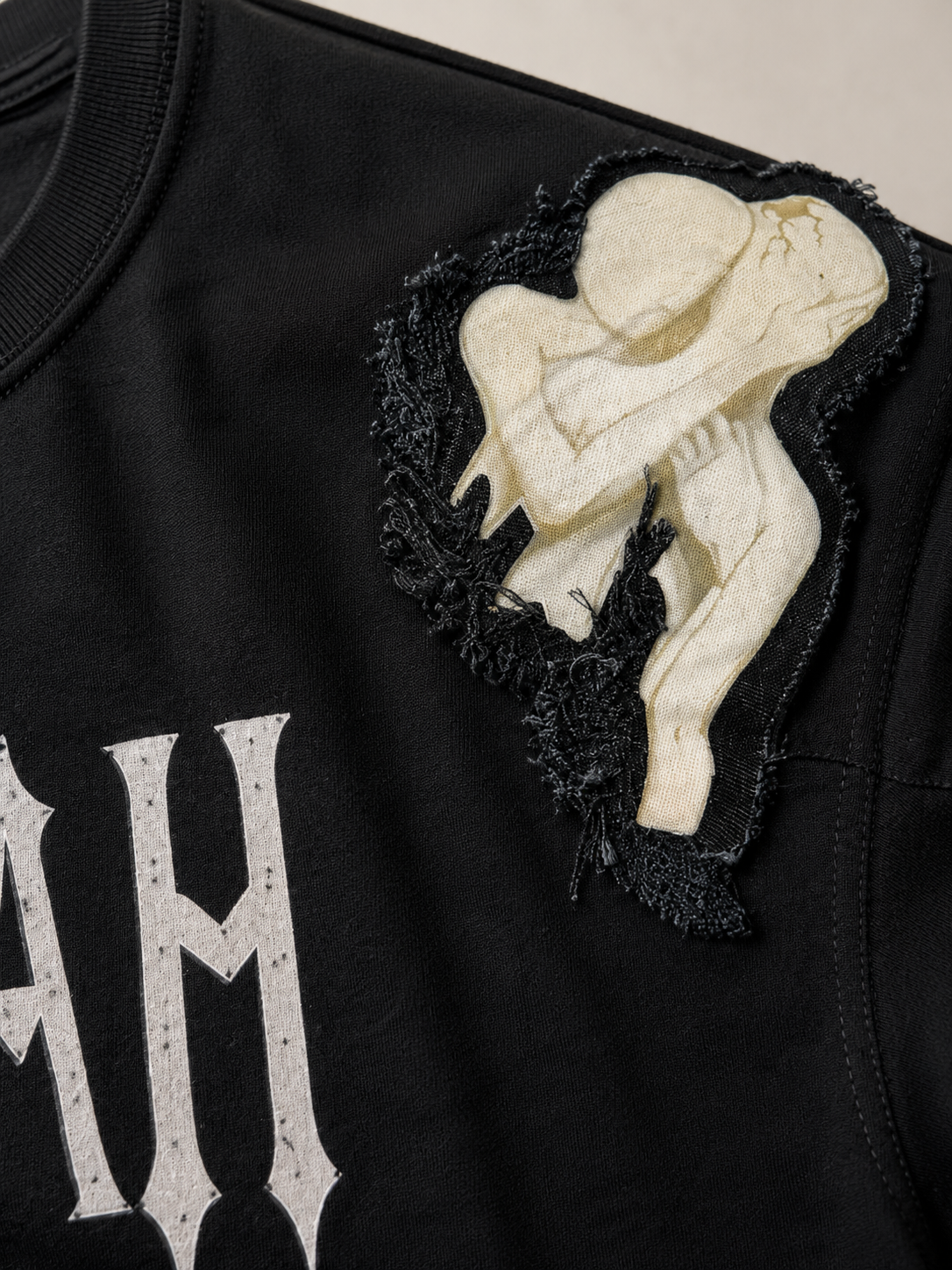 Close-up of a black garment featuring a large cream-colored skeleton figure applique on the shoulder with black lace trim, alongside a distressed white graphic lettering.