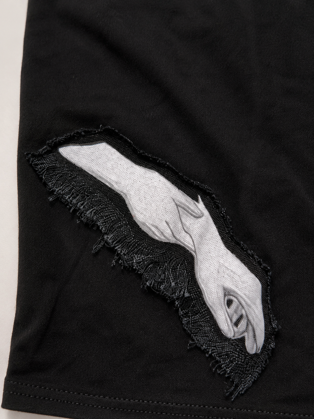 Close-up of a black garment with a distressed ripped section showing a white embroidered hand graphic.