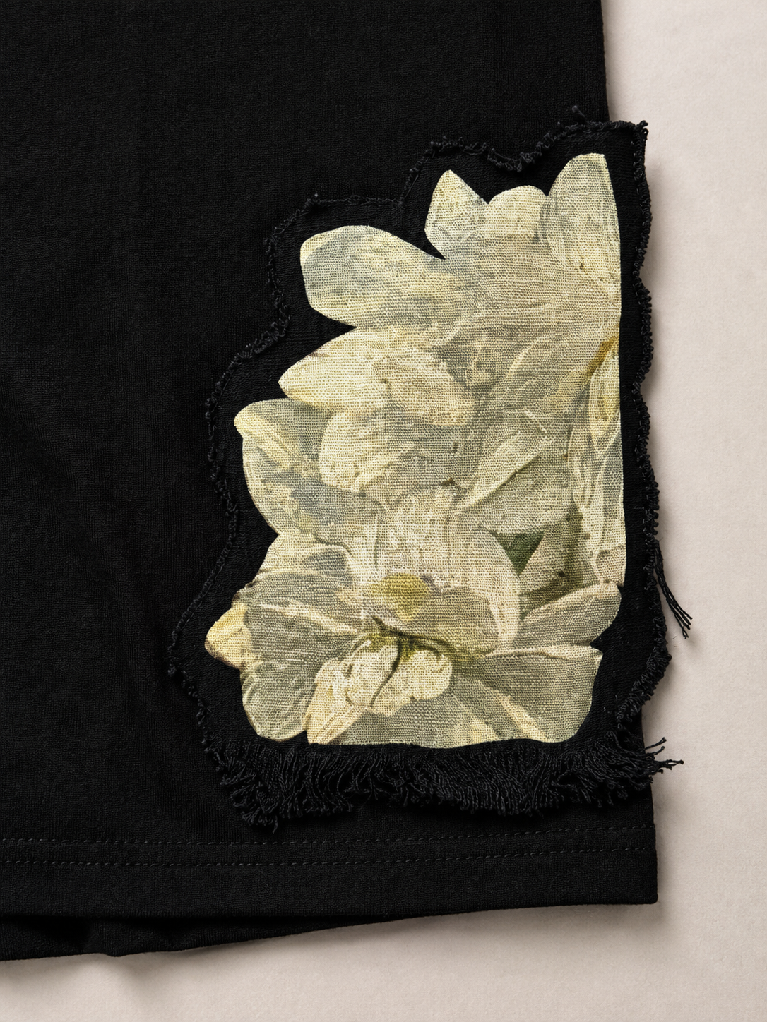 A close-up of a black garment featuring a large, distressed applique patch of pale yellow and cream textured fabric flowers with frayed edges.