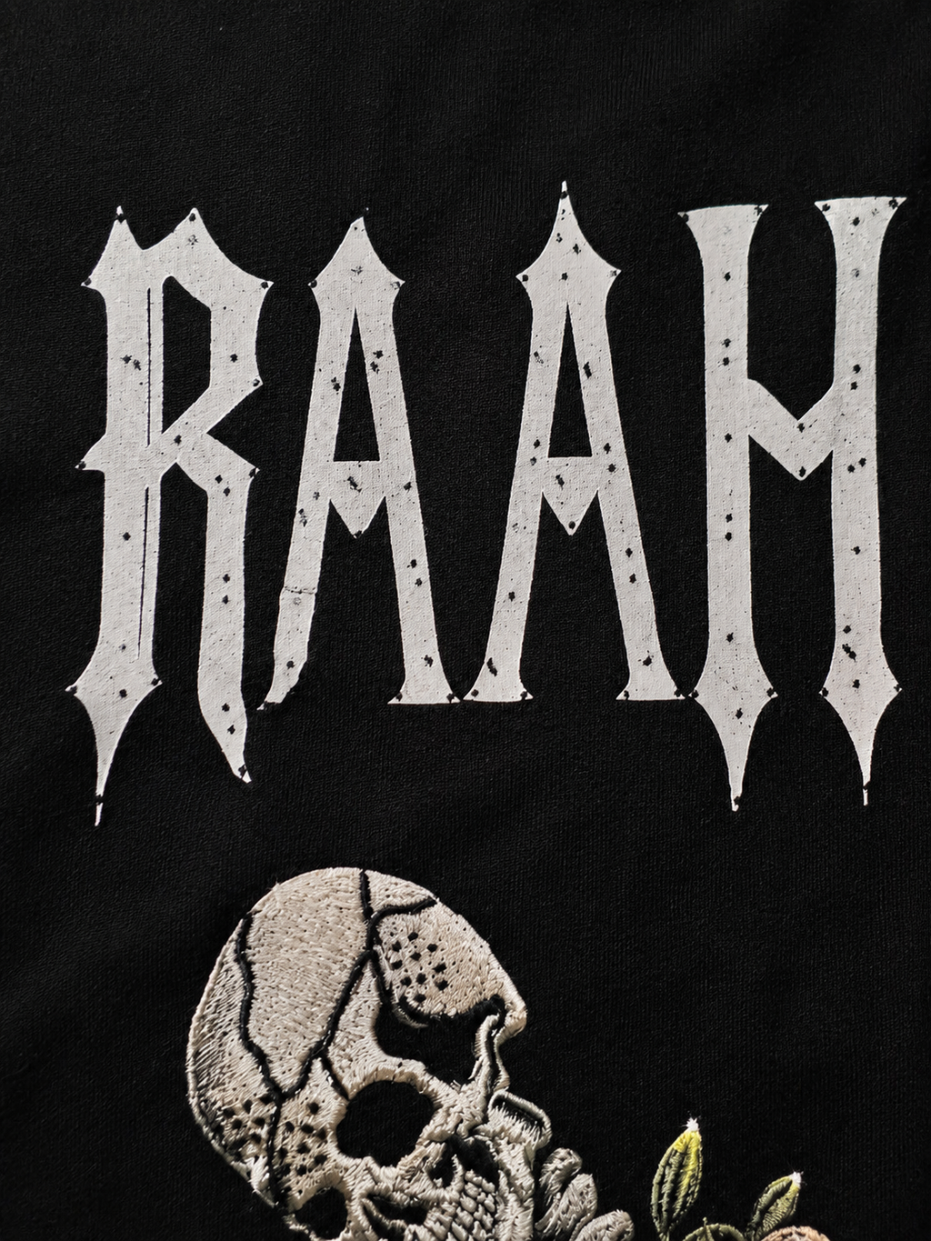 Close-up of a black garment featuring a large white gothic-style 'RAAH' graphic above a detailed skull illustration with small green leaf accents.