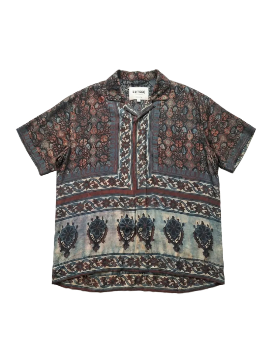 A short-sleeve button-up shirt with a complex batik-style floral and geometric pattern in dark brown, beige, and muted blue tones, laid flat against a white background.