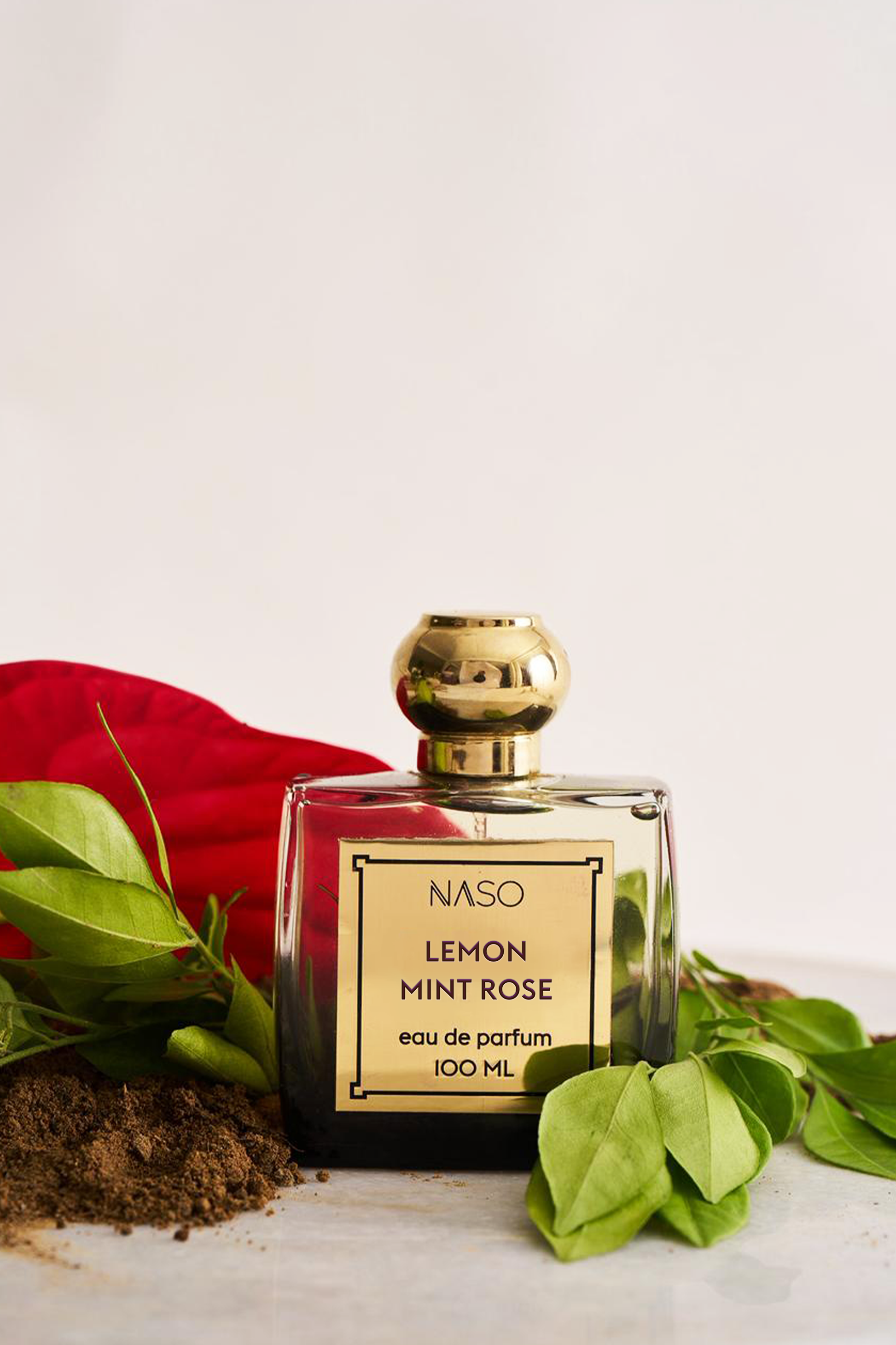 A 100ml bottle of NASO Lemon Mint Rose Eau de Parfum, featuring a clear glass body, dark liquid, a gold cap, and a gold label. The bottle is styled on a light surface with green mint leaves, brown powder, and a red fabric prop against a clean white background.