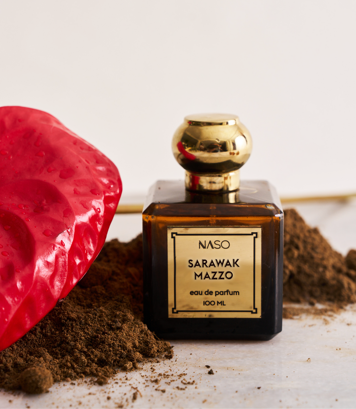 A bottle of NASO Sarawak Mazzo Eau de Parfum with a gold cap and gold label, styled on a white surface with brown powder and a red textured stone prop against a light background.