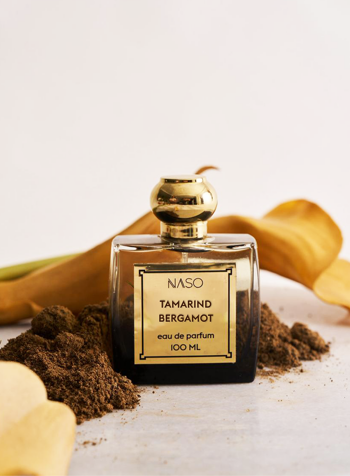 A 100ml bottle of NASO Tamarind Bergamot Eau de Parfum, featuring a clear glass body, a gold cap, and a gold label with black text. The bottle is styled on a white surface with scattered tamarind powder and dried yellow tamarind peel in the background.
